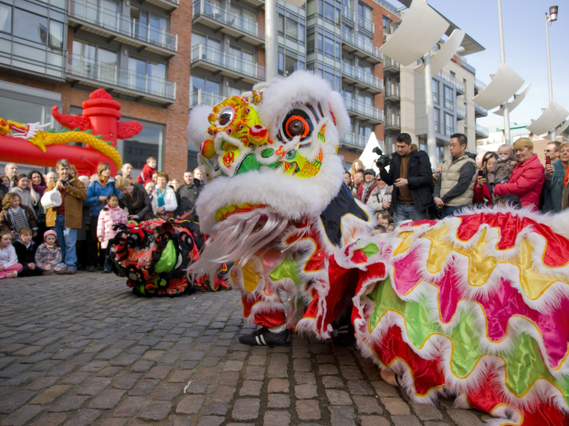 Ein lebendiges chinesisches Neujahrsfest in Amsterdam mit einer Löwen-Tanz-Show vor einer Zuschauermenge, einige halten Kameras, vor einem Hintergrund aus Gebäuden, Laternenmasten und einem klaren blauen Himmel.
