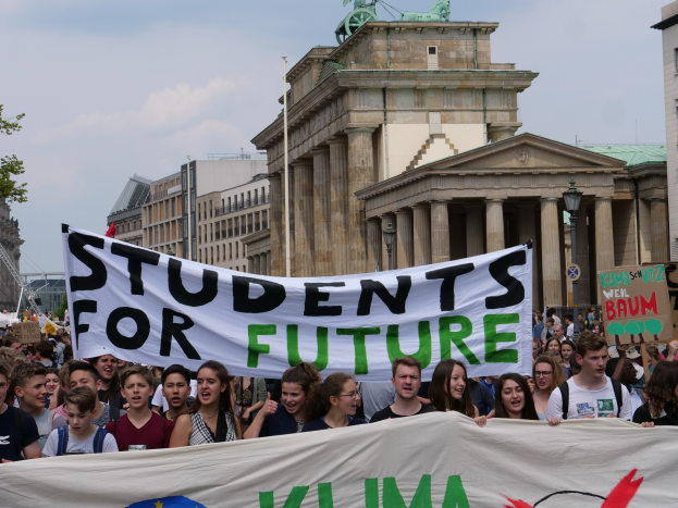 Eine Gruppe von Schülern marschiert in Berlin, eine buntfarbene Fahne mit der Aufschrift "Students for Future" schwingend, mit Gebäuden, Bäumen und Himmel im Hintergrund.