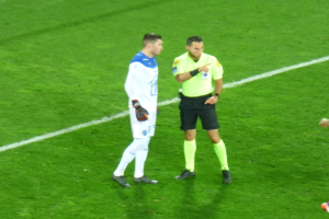 Schiedsrichter in gelbem Hemd und schwarzen Shorts, der mit einem Fußballspieler in weiß-blauer Uniform auf dem Feld spricht, beide tragen Turnschuhe.