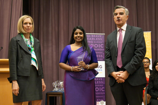 Zwei Frauen und ein Mann stehen auf einer Bühne mit einem braunen Vorhang dahinter; die Frau in der Mitte hält einen Award, und mehrere Awards sind auf dem Tisch vor ihnen.