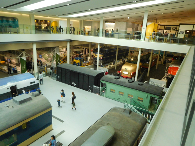 Ein großer Museumsraum mit verschiedenen Arten von Zügen, begleitet von Menschen, Geländern, Säulen, Lampen und Informationsschildern.