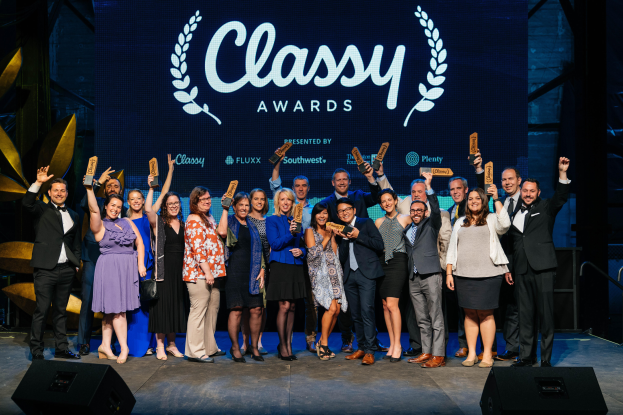 Eine Gruppe von Menschen auf einer Bühne, die lächeln und Auszeichnungen halten, mit Lautsprechern unten und einem Hintergrundbild mit 'Classy Awards 2019'.