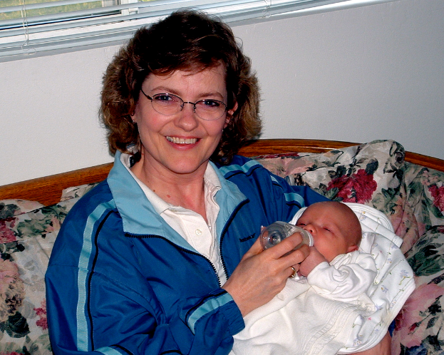 Eine Frau in einer blauen Jacke sitzt auf einem Sofa und f├╝ttert ein Baby in der N├Ąhe eines Fensters mit Jalousien.