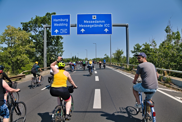 Eine Gruppe von Menschen in Helmen fährt auf Fahrrädern eine Straße entlang, mit einem Geländer auf einer Seite und Bäumen auf der anderen, unter einem klaren blauen Himmel mit Laternenmasten und einer Texttafel oben.