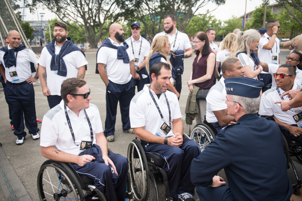 Eine Gruppe von Menschen im Rollstuhl unterhält sich vor einer Kulisse aus Bäumen, Pfählen, Gebäuden und einem klaren blauen Himmel, wobei einige Mützen und Schutzbrillen tragen, was darauf hindeutet, dass sie bei den Paralympischen Spielen sind.