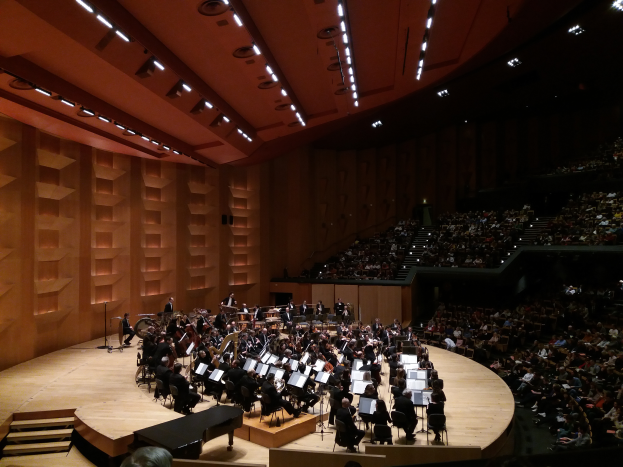 Ein großer Saal voller Menschen, die ein Konzert in der Royal Albert Hall in London besuchen, einige sitzen und halten Musikinstrumente, ein paar stehen auf der Bühne, Lampen an der Decke und eine Wand auf der linken Seite.