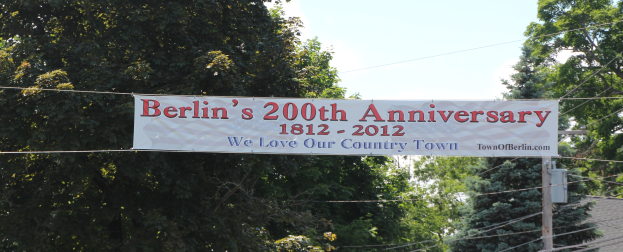 Ein Banner mit der Aufschrift 'Berliner Jubiläum 200' hängt an einem Draht in der Mitte, mit Bäumen im Hintergrund und einem Haus auf der rechten Seite, unter einem sichtbaren Himmel.