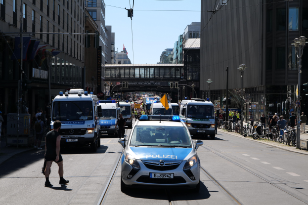 Polizeiautos fahren eine Straße mit hohen Gebäuden, Fußgängern auf dem Gehweg, parkenden Fahrrädern, Laternen, einer Brücke im Hintergrund und einem klaren blauen Himmel.