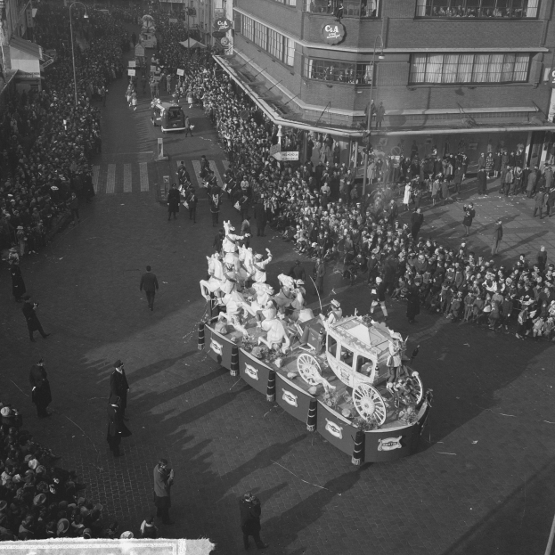 Eine große Menschenmenge umringt einen Festwagen bei einem Umzug, der mit bunten Luftballons und Bändern geschmückt ist, mit Gebäuden und Laternen im Hintergrund, in Schwarz-Weiß.
