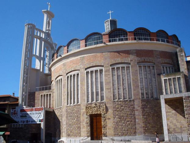 Die Kirche des Heiligen Grabes, ein großes Gebäude mit einem zentralen Uhrenturm, umgeben von Fenstern, einer Tür, einer Fahne, einem Metallzaun, einer Gruppe von Menschen, Fahrzeugen, Zelten und einem bewölkten Himmel.