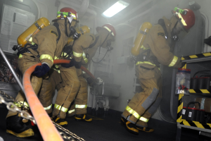Feuerwehrleute in Schutzbekleidung und Helmen arbeiten an einem Feuerlöschwagen mit Sauerstoffflaschen, während ein Gestell mit Gegenständen und im Hintergrund Rohre, Lampen und andere Gegenstände zu sehen sind.