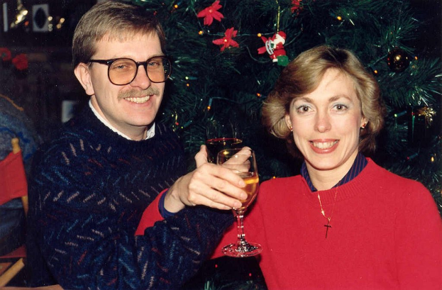 Ein Paar lächelt, whährend es Gläser mit Wein hält, mit einem Weihnachtsbaum im Hintergrund.