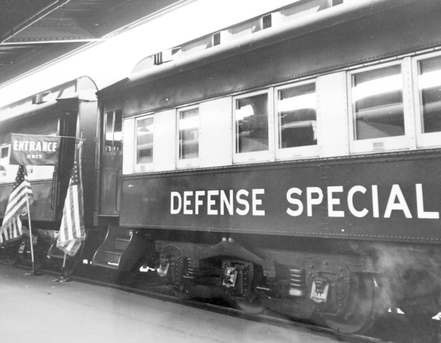 Schwarzes und weißes Foto eines Zuges mit der Aufschrift "Defense Special" auf den Eisenbahnschienen mit zwei Fahnen links und einer Decke darüber.