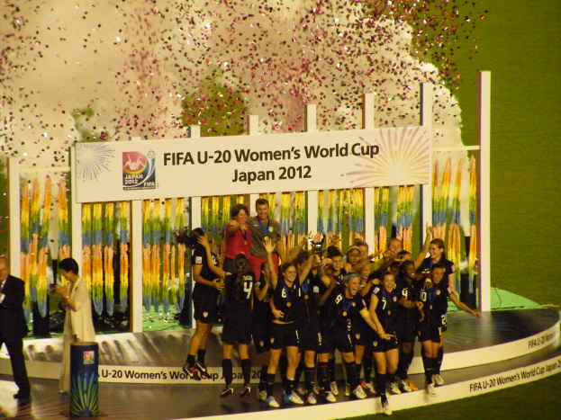Eine Gruppe von Fussballspielerinnen in schwarzen Uniformen steht auf einer Bühne und feiert ihren Sieg, umgeben von einer Menge mit Kameras, mit einem dekorierten Hintergrund und einem grasbewachsenen Boden.