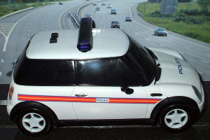 Ein weißer Polizeiwagen mit der Aufschrift "Polizei" fährt auf einer Straße außerhalb einer Stadt, umgeben von Gras, Bäumen und anderen fahrenden Fahrzeugen.