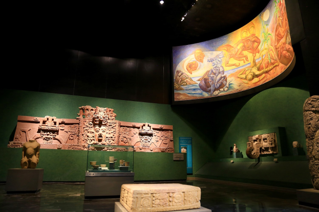 Ein Museumsraum mit verschiedenen Artefakten, darunter Skulpturen, Gegenstände auf Tischen, eine Texttafel, eine Wandmalerei und Deckenleuchten im Anthropologischen Museum von Mexiko-Stadt.