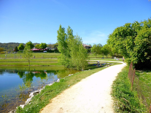Ein Schotterweg neben einem Gew√§sser, umgeben von gr√ľner Vegetation und einem Zaun, mit Menschen, die die Landschaft genie√üen, Geb√§uden, H√∂llen und einem klaren blauen Himmel im Hintergrund, der einen idealen Schauplatz f√ľr eine Fahrradtour in Frankreich bildet.