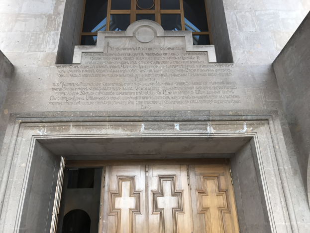Große hölzerne Tür vor einem Steinbau mit einem Glasfenster oben; Inschrift auf der Wand erinnert wahrscheinlich an Holocaust-Opfer.