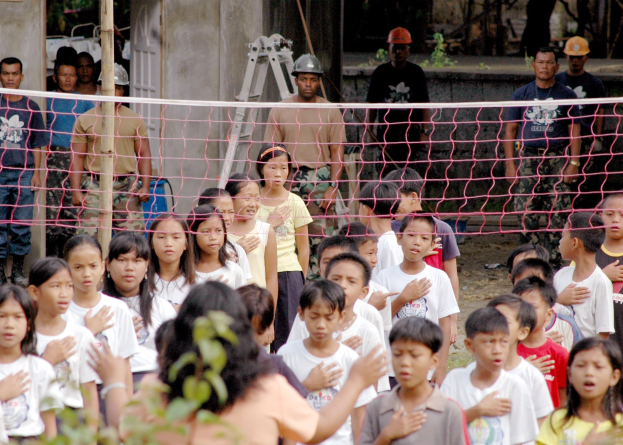 Kinder und eine Frau neben einem Netz mit Helmträgern, einer Leiter, einer Wand und Bäumen im Hintergrund.