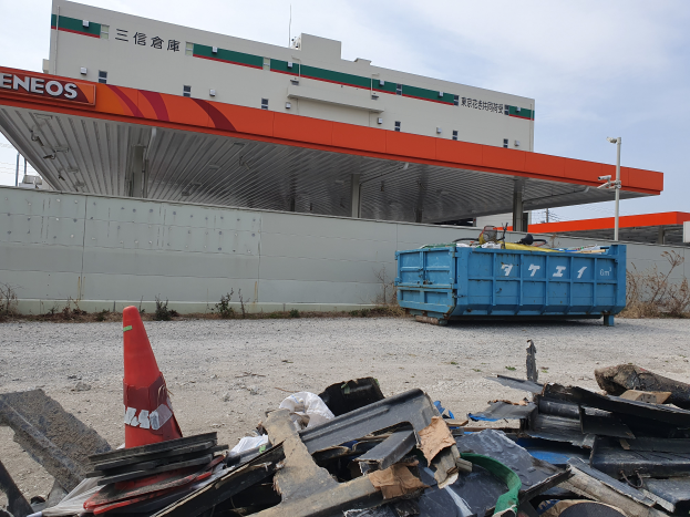 Ein großes Gebäude mit erheblichen Trümmern, darunter Schrott, ein Verkehrskegel und ein blauer Container davor, mit Pflanzen, Polen und dem Himmel im Hintergrund, was auf Abrissarbeiten hindeutet.