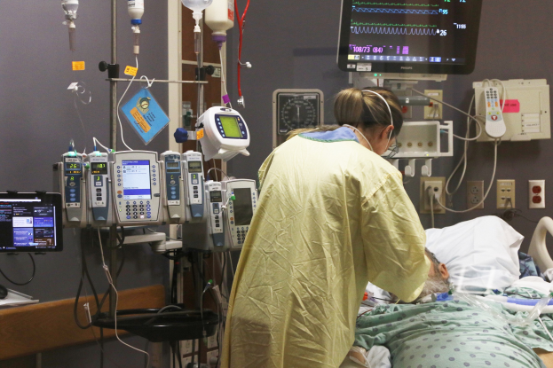 Ein Mann liegt in einem Krankenhausbett neben einem Monitor, mit einer Frau, die neben ihm steht; ein Tisch mit einem weiteren Monitor ist links und medizinische Geräte sind im Hintergrund zu sehen.