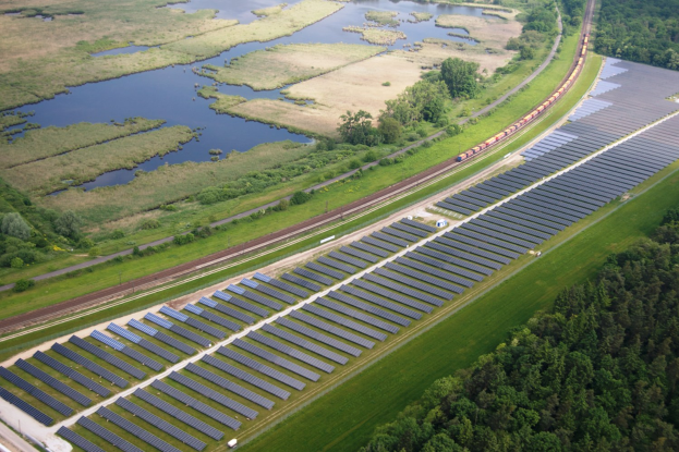 Luftaufnahme einer Solarpark mit Panelen, umgeben von B"umen, Gras, Wasser und einer Bahn auf einer Schiene.
