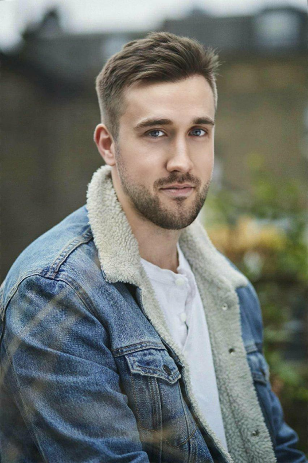 Ein bärtiger Mann in einer Jeansjacke und einem weißen T-Shirt steht vor einem unscharfen Hintergrund und präsentiert einen stylischen Kurzhaarschnitt.