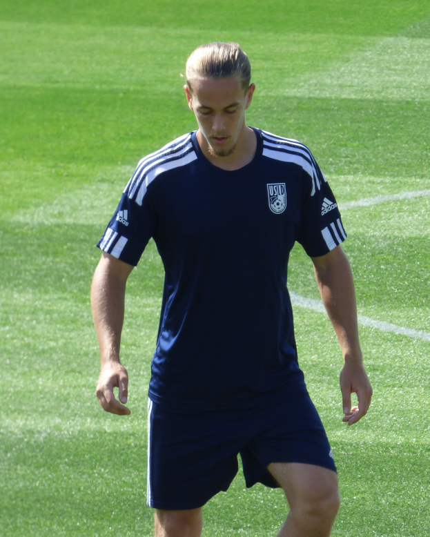 Ein Mann in einem blauen T-Shirt und Shorts geht selbstbewusst über ein grünes Feld und sieht aus wie ein professioneller Füßballer mit entschlossener Miene.
