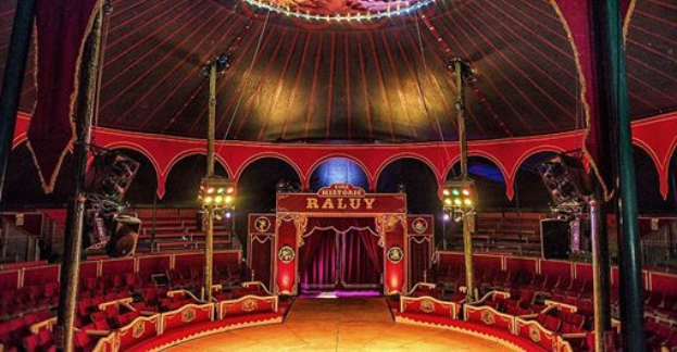 Das Innere eines großen Zirkuszeltes mit roten Sitzplätzen, Stangen, Lichtern, einer Bühne, einem Vorhang, einem Namensschild und anderen Gegenständen, wahrscheinlich das historische Raluy Theatre.