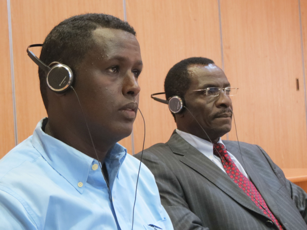 Zwei Männer in einem Gerichtssaal mit Kopfhörern, einer in einem blauen Hemd links und einer mit Brille in der Mitte, mit einer hölzernen Wand im Hintergrund.
