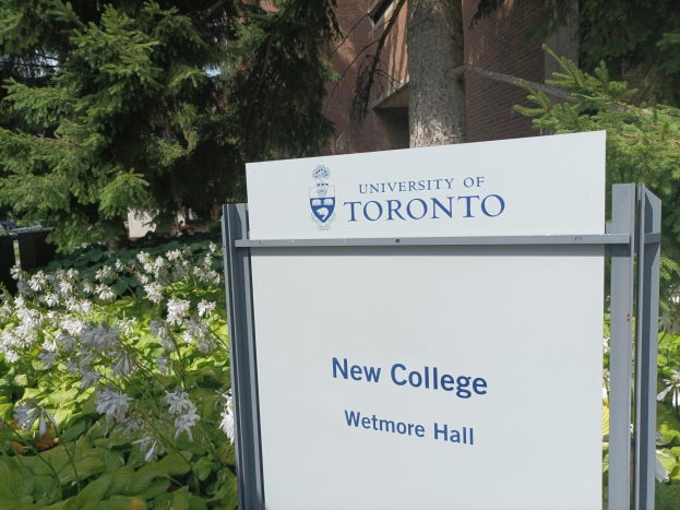 Außenansicht des neuen Colleges der Universität Toronto an Wetmore Hall mit einem Schild vor dem Gebäude, umgeben von grünem Gras, bunten Blumen, hohen Bäumen und einem klaren blauen Himmel.