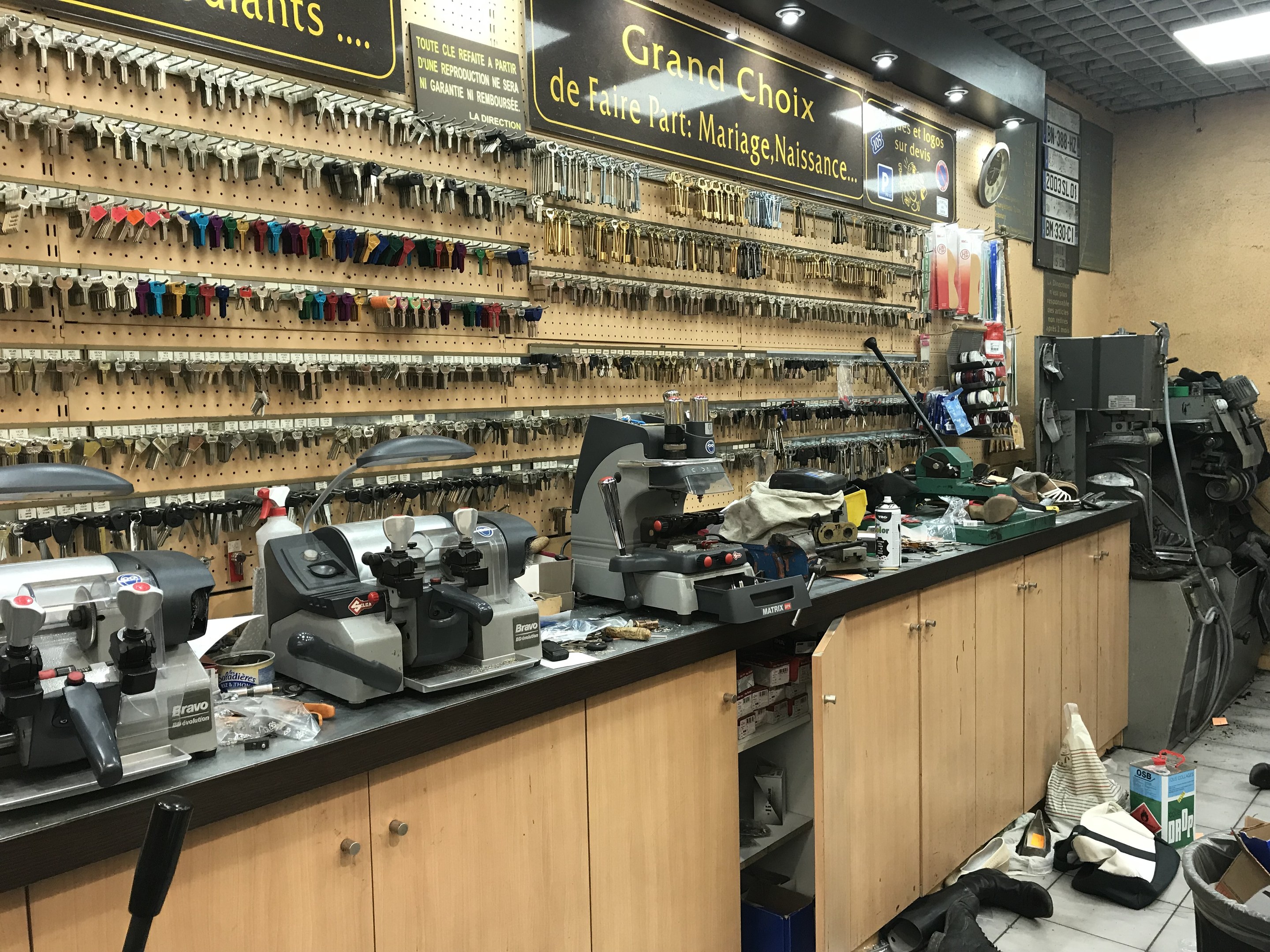 Ein unordentlicher Garage-Workshop mit verschiedenen Werkzeugen, Maschinen, Schränken und Tafeln an den Wänden und auf dem Boden, eine Uhr und Deckenleuchten.