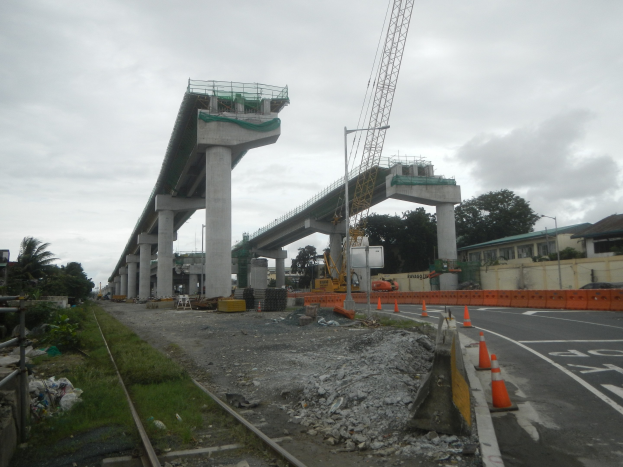 Baustelle mit einer Brücke im Hintergrund, Straße mit Absperrgittern, verstreute Steine und Gras, Eisenbahnschienen auf der linken Seite, Bäume und Gebäude säumen die Straße und ein bewölkter Himmel.