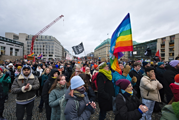 Große Menschenmenge bei einer LGBTQ+-Rechtsdemo in Berlin, mit Fahnen und Plakaten, im Hintergrund Gebäude, ein Kran und ein bewölkter Himmel.
