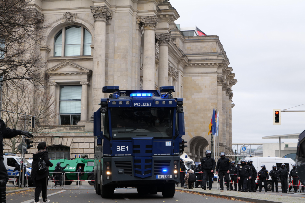 Gruppe von Polizeibeamten vor einem großen Gebäude mit Fenstern, Säulen und Bögen, mit Fahrzeugen, einer Person mit einer Kamera, Bäumen, Verkehrszeichen, Flaggen und einem klaren blauen Himmel.