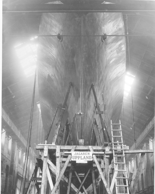 Schwarzes und weißes Foto eines großen Boots im Bau, das eine Leiter, Holzpfosten, ein Schild, Metallrahmen, Deckenleuchten und Fenster zeigt.