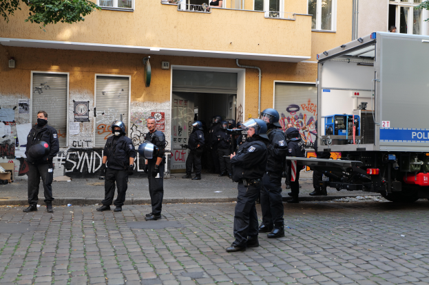 Eine Gruppe von Polizisten in Helmen, die Waffen halten, steht vor einem Gebäude mit Fenstern, Rollläden und Graffiti, mit einem Truck in der Nähe und Bäumen im Hintergrund.