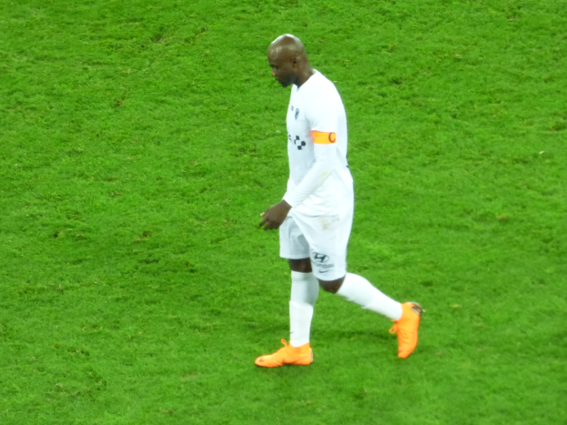 Ein Fussballspieler in einem weissen T-Shirt, weissen Shorts und orangefarbenen Schuhen läuft über ein saftiges grünes Feld.