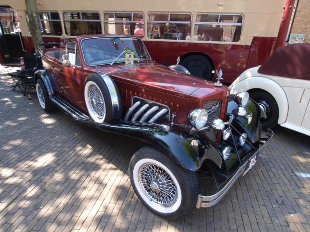Ein Vintage-Auto neben einem Doppeldeckerbus auf einer Kopfsteinpflasterstraße geparkt, mit Stühlen und einem Baum im Vordergrund, einem Gebäude im Hintergrund und Menschen im Bus sichtbar.