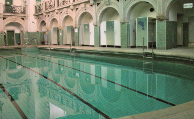 Innenbad mit klarem Wasser, umgeben von Fliesenboden, Bogengängen, Säulen, Geländern, Türen und Texttafeln in einem großeen Gebäude.