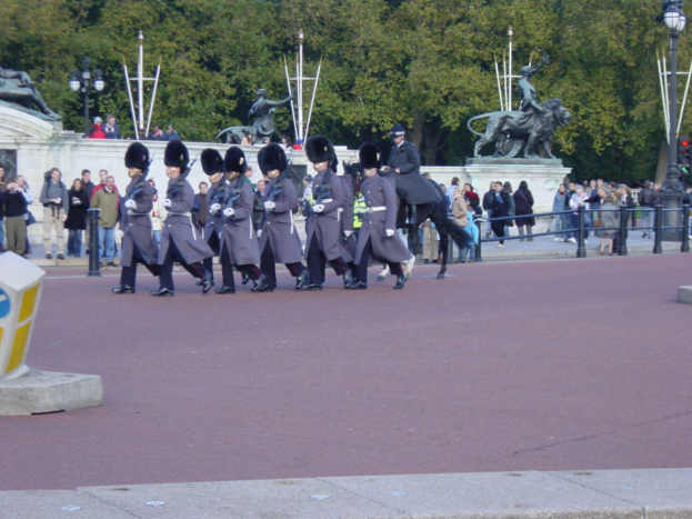 Eine Gruppe von Soldaten in grauen Uniformen marschiert mit Gewehren, ein Mann zu Pferd neben ihnen, Zuschauer schauen zu, Statuen und Bäume im Hintergrund.