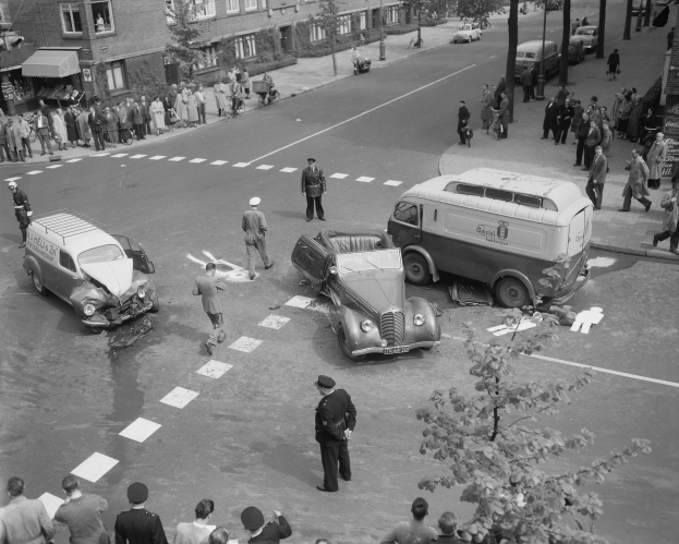 Eine Schwarz-Weiß-Szene eines Autounfalls an einer Straßenecke mit mehreren Fahrzeugen, darunter ein Lieferwagen, einer Gruppe von Menschen drumherum und im Hintergrund Gebäude, Bäume, Masten und Texttafeln.