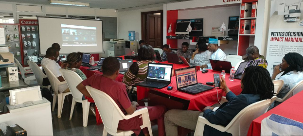 Eine Gruppe von Menschen sitzt um einen Tisch mit Laptops, Mäusen, Flaschen und anderen Gegenständen und nimmt an einem digitalen Marketing-Workshop teil, wobei ein Banner, ein Display-Bildschirm, eine Tür und elektronische Geräte im Hintergrund zu sehen sind.