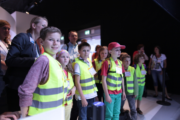 Eine Gruppe von Kindern trägt Sicherheitswesten und Namensschilder und steht auf einem Boden, wobei ein Junge links lächelt und ein Pfahl rechts steht, während im Hintergrund ein paar Menschen stehen.