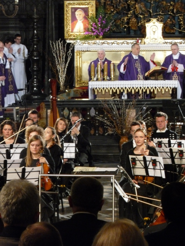 Eine Gruppe von Menschen, die Geigen spielen, mit Noten vor sich, mit drei Kirchenvätern im Hintergrund, einer Bibel vor ihnen und einem Rahmen mit Schnitzereien dahinter.