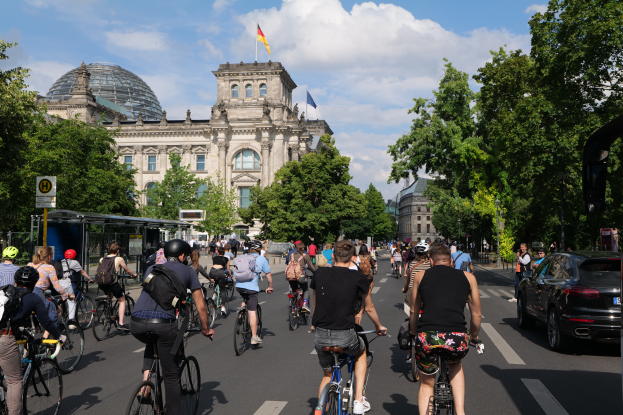 Eine Gruppe von Menschen, die auf Fahrr√§dern eine stra√če in Berlin, Deutschland, entlangfahren, mit B√§umen auf beiden Seiten, einem Bushaltestellen auf der rechten Seite und einer Flagge, die auf einem der Geb√§ude weht, unter einem bew√∂lkten Himmel.
