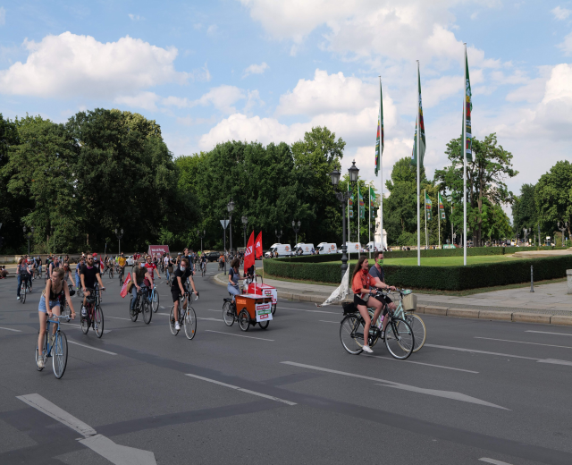 Eine Gruppe von Menschen, die auf Fahrrädern eine von Bäumen gesäumte Straße während der Paris Bike Ride 2016 hinunterfahren, mit Laternenmasten, Fahnenmasten, bewölktem Himmel und ein paar Fahrzeugen im Hintergrund.
