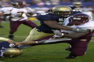 Zwei Fußballspieler in Sportkleidung und Helmen auf dem Feld zusammenstoßen, während andere im Hintergrund zuschauen.