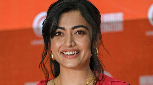 Eine Frau in einem roten Kleid mit einem eleganten Dutt und dezentem Make-up lächelt in die Kamera, trägt schwarze High Heels und steht vor einem hellgelben Banner mit der Aufschrift "Sonam Kapoors beste Schönheits Looks vom Cannes Film Festival."