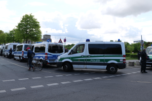 Eine Gruppe von Polizeiwagen entlang einer Straße geparkt mit einem Radfahrer im Vordergrund und Gebäuden, Bäumen und Schildern im Hintergrund unter einem bewölkten Himmel.